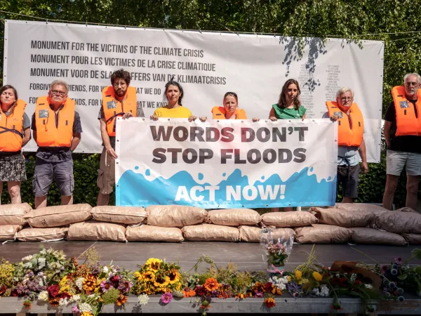 Aktion zum EU-Gedenktag für die Opfer der globalen Klimakrise im Juli 2026 in Brüssel: Menschen in orangefarbenen Rettungswesten halten Transparent mit der Aufschrift "Words don´t stop floods. Act Now!"